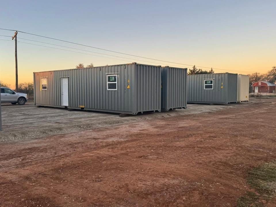 Two shipping-container housing units on a graded gravel pad at sunset